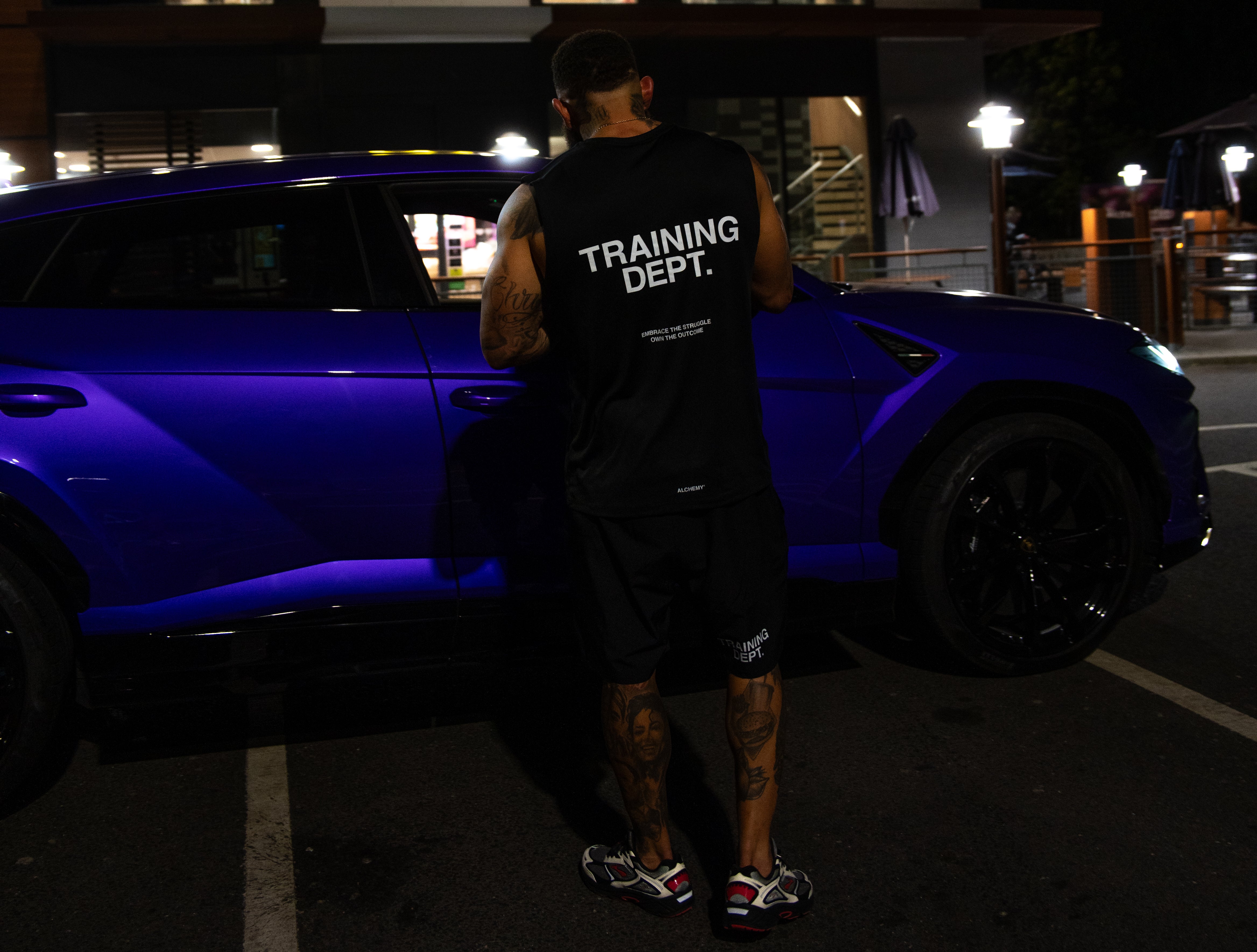 Person wearing a 'Training Dept.' shirt standing next to a purple car at night.